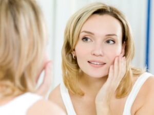 woman in white top looks at her face in the mirror